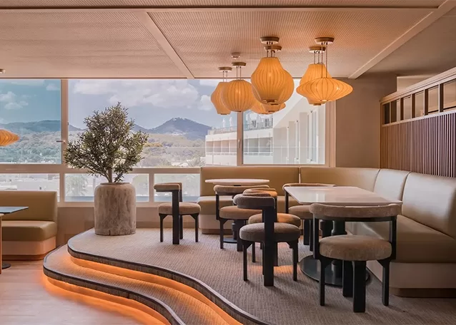 A modern, stylish lounge with tiered seating and a large potted plant. Pendant lights hang from the ceiling, and large windows offer a scenic view of mountains. Soft lighting creates a cozy ambiance.