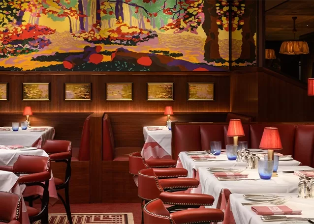 A cosy restaurant with red leather booths, white tablecloths, and red lamps on tables. The walls are adorned with colorful landscape art and framed paintings, creating a warm and inviting atmosphere.