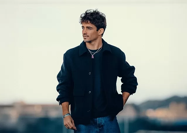 Charles Leclerc - with curly hair stands outdoors wearing a dark jacket, black shirt, and jeans. He has a thoughtful expression, with his right hand in his pocket. The background is blurred, showing a soft, neutral sky and some indistinct structures.