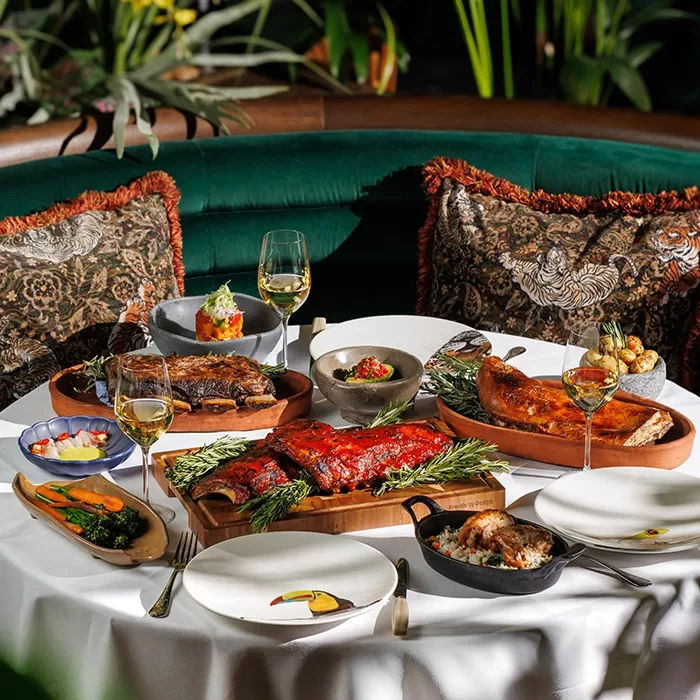 Elegant restaurant table set with various gourmet dishes, including ribs, baked vegetables, bread, and wine glasses, surrounded by lush plants and plush, patterned cushions on a green velvet booth.