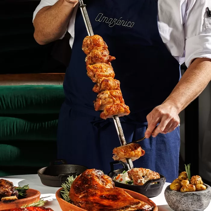 A chef wearing a blue apron with Amazónico embroidered carves pieces of grilled meat from a large skewer onto a plate, surrounded by various dishes of roasted meats and vegetables.