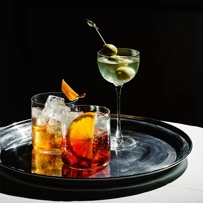 A silver tray holds three cocktails: a martini with green olives on a pick, an amber drink with ice and an orange twist, and a red cocktail with ice and a large orange slice garnish, all against a dark background.