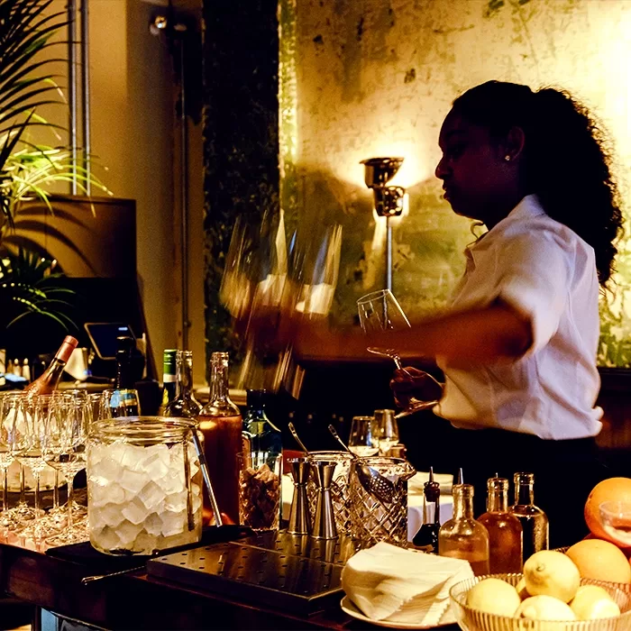 A bartender in a white shirt prepares drinks behind a bar, surrounded by glasses, bottles, ice, and fresh lemons, with warm ambient lighting and a textured wall in the background.