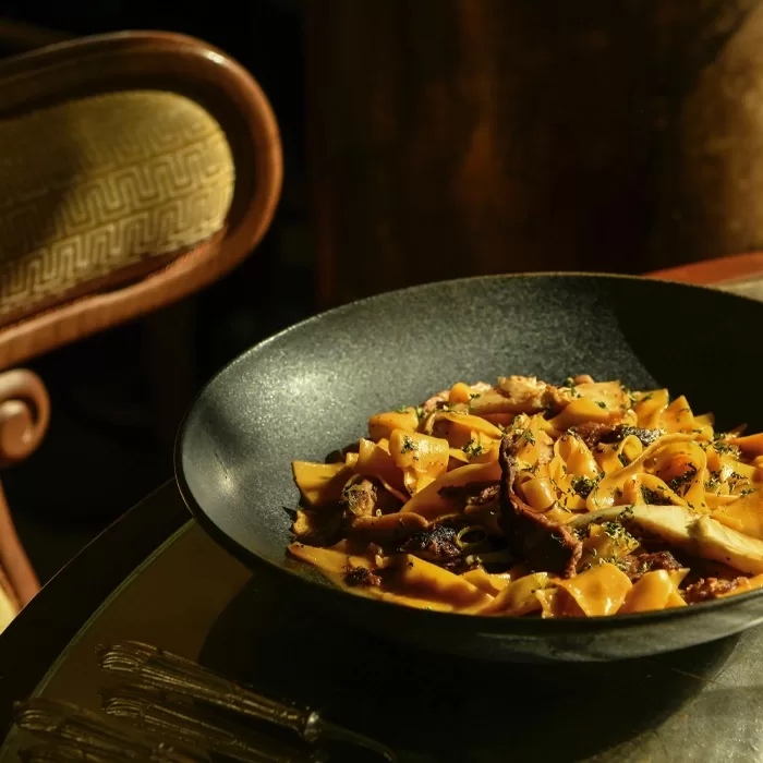 A bowl of pasta with mushroom slices and herbs sits on a dark plate atop a glass table, with a vintage upholstered chair in the background, lit by warm, natural light.