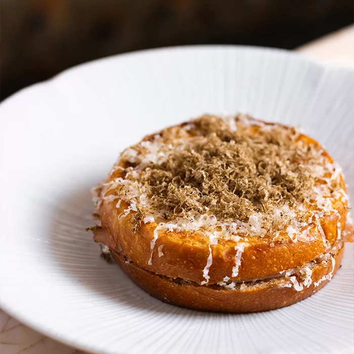 A golden, toasted sandwich topped with finely grated cheese and shaved truffle sits on a white, textured plate.