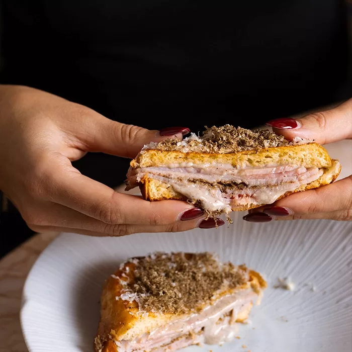 A person with dark red nails holds a half-eaten sandwich filled with layers of ham and melted cheese. The sandwich is sprinkled with grated truffle and sits above a white plate with another half on it.