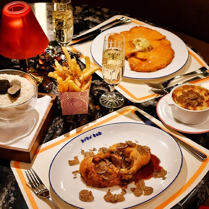 A table set with elegant dishes, including roast chicken with truffles, fries in a cup, a bowl of creamy gratin, breaded cutlets, glasses of sparkling wine, and a lamp, all on stylish placemats.