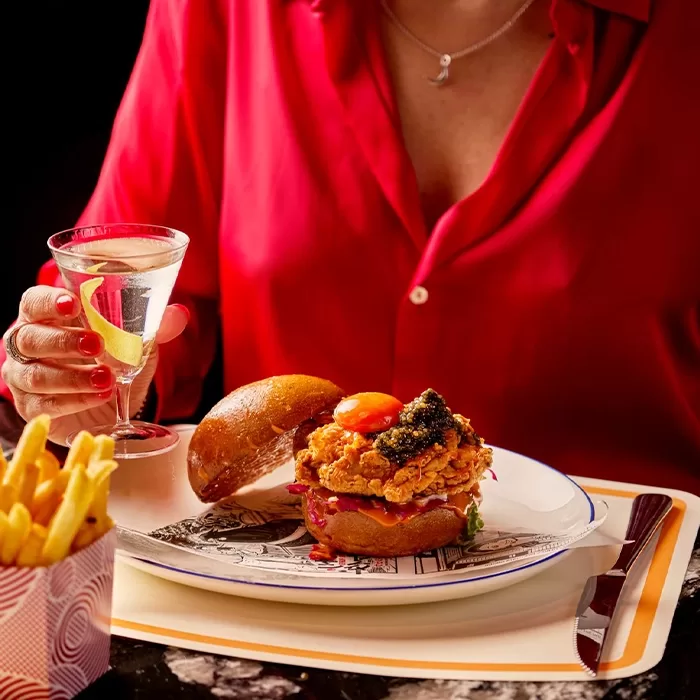 A person in a red shirt holds a martini with a lemon twist, seated at a table with a fried chicken sandwich topped with caviar, a side of fries, and a placemat with a graphic design.