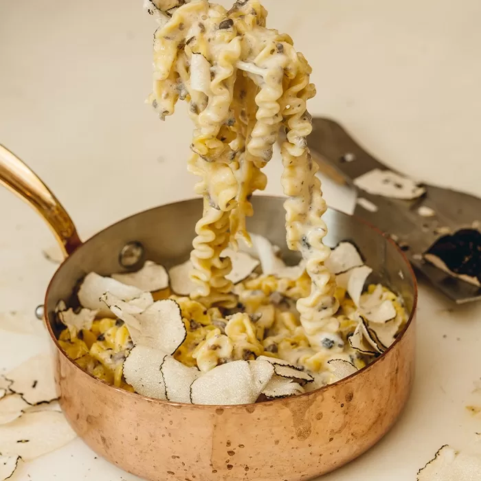 A copper pan filled with creamy pasta, topped with thin slices of white truffle. A fork lifts a portion of the pasta, showing its rich, cheesy sauce and truffle garnish. A truffle slicer rests nearby.