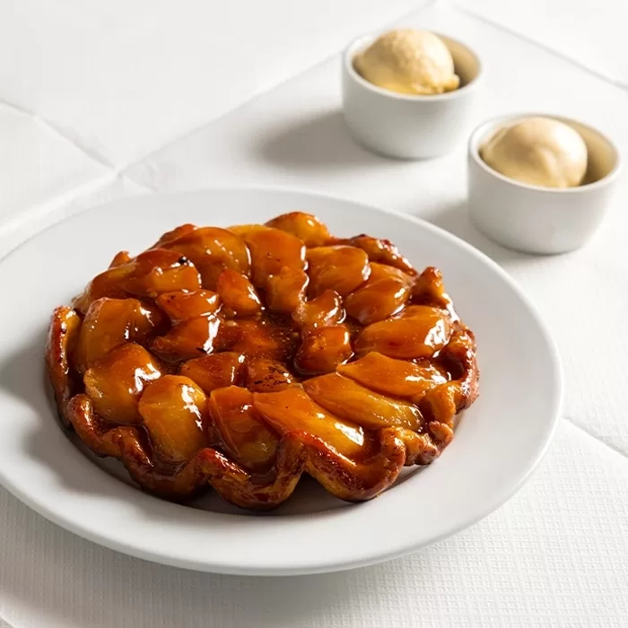 A glazed apple tart sits on a white plate, with two scoops of vanilla ice cream in small bowls in the background on a white tablecloth.