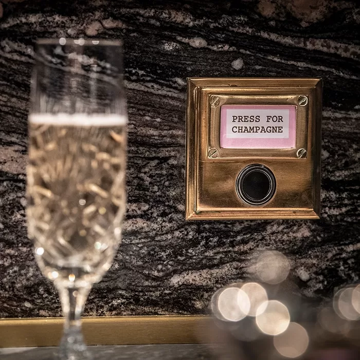 A glass of champagne stands in front of a marble wall with a gold button panel labeled Press for Champagne.