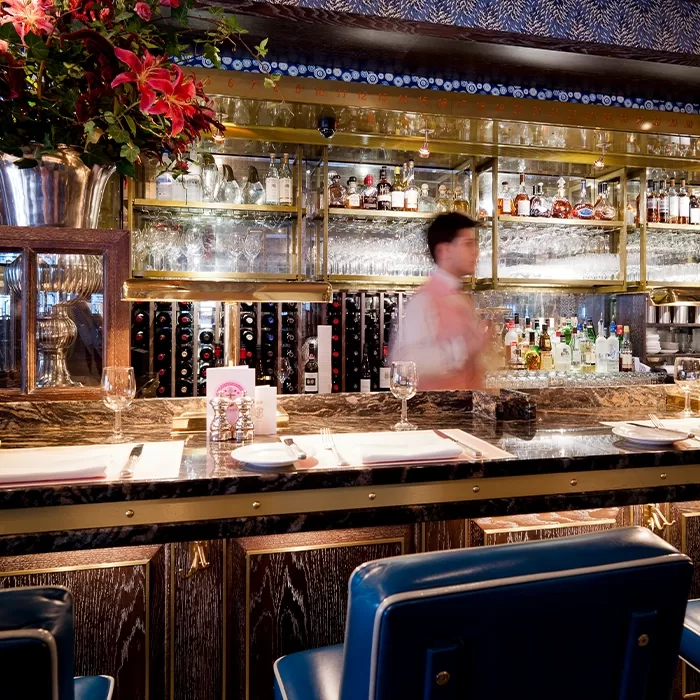 A bartender in a white shirt moves behind a marble bar with blue chairs, wine glasses, and place settings. Shelves with bottles, glassware, and flowers decorate the background.