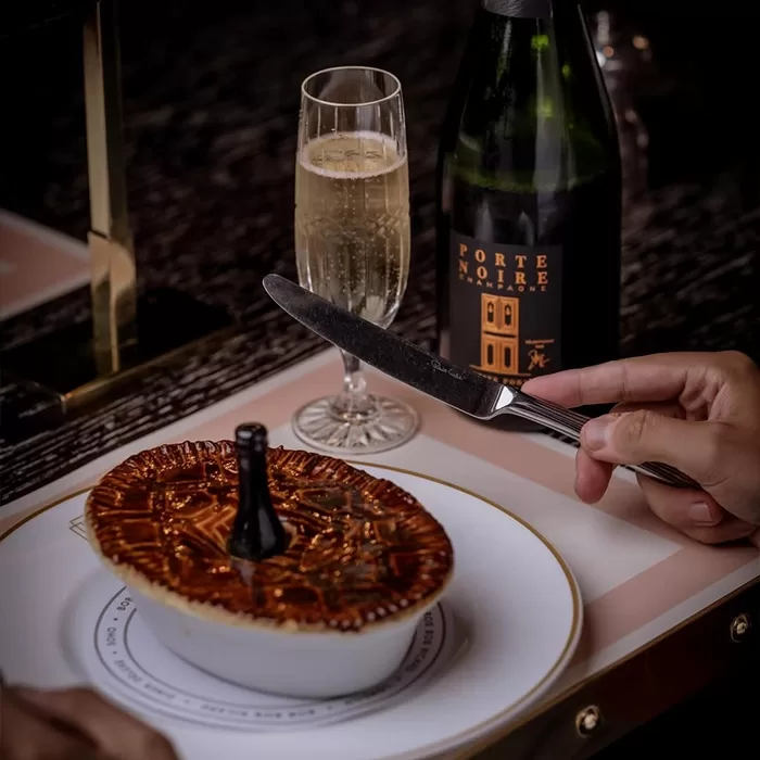 A hand holds a knife above a golden-brown pastry pie topped with a small wine bottle, served on a white plate. Beside it are a champagne flute filled with sparkling wine and a dark bottle labeled Porte Noire.