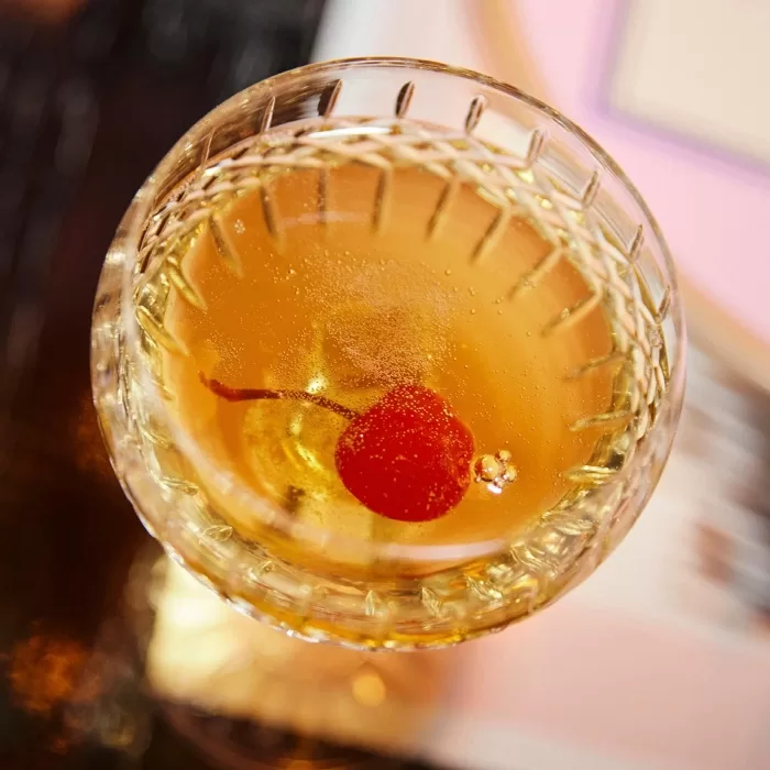 A close-up, top view of a clear cocktail in a patterned glass, garnished with a red cherry at the bottom.