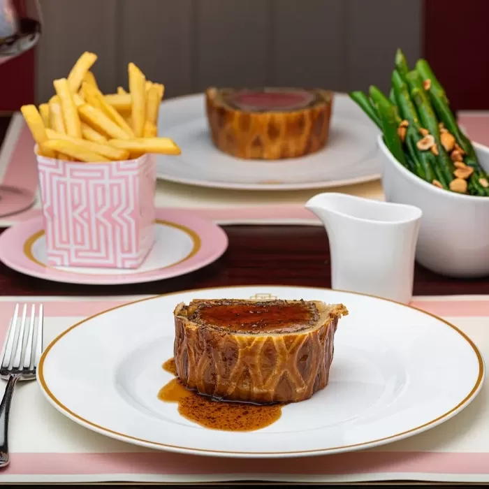 A plate with a slice of beef Wellington topped with sauce, accompanied by a cup of French fries and a side of green beans with nuts, set on a restaurant table with elegant tableware.