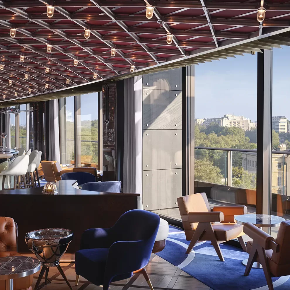 Modern interior lounge with large windows offering a city view. The space features contemporary furniture, including armchairs and tables, under a ceiling with string lights. Natural light fills the room, highlighting the blue and brown color scheme.