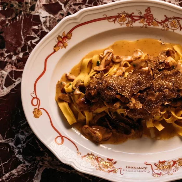A white ornate plate with tagliatelle pasta topped with creamy mushroom sauce and generous shavings of black truffle, set on a dark marbled table.