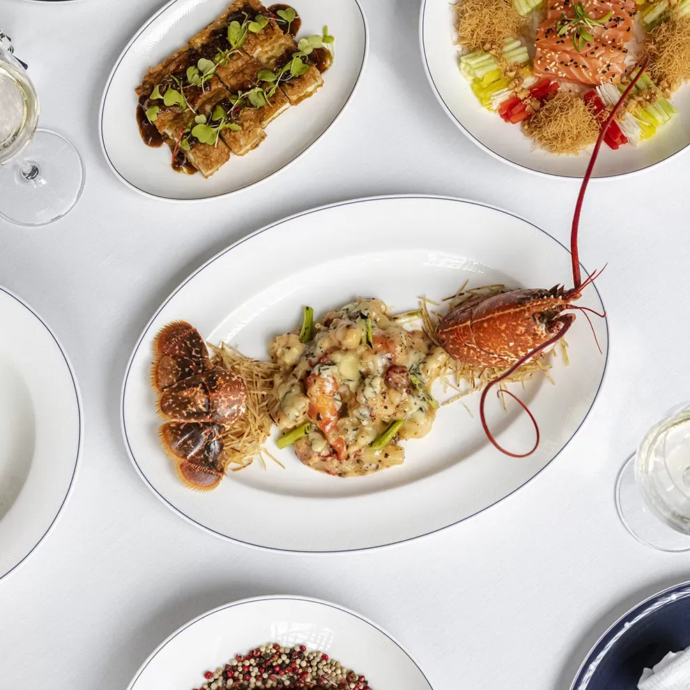 A white tablecloth displays an elegant seafood dish with lobster and garnishes on an oval plate. Surrounding dishes include what appears to be salmon and other gourmet offerings, alongside wine glasses and a dish of peppercorns.