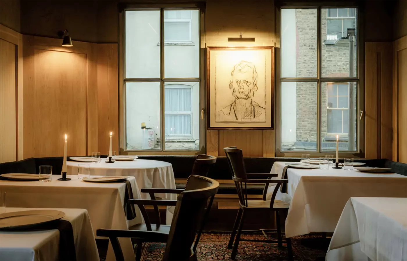 A cosy restaurant interior features tables with white tablecloths and lit candles. Soft lighting from a window illuminates a monochrome portrait on the wall, creating an intimate ambiance at Cycene, Shoreditch.