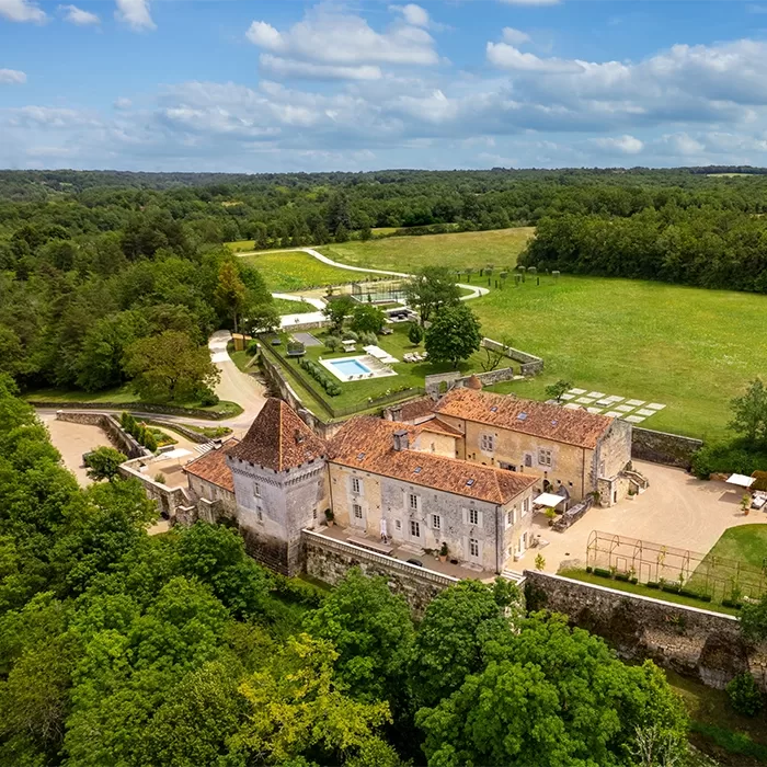 Aerial view of Château de Chanet - a large, historic chateau with a red-tiled roof surrounded by lush greenery. A swimming pool and manicured gardens are visible nearby. The landscape includes expansive lawns and a wooded area under a partly cloudy sky.