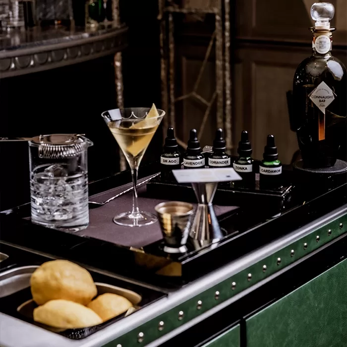 A cocktail station with a martini glass, mixing glass with ice, droppers of flavor extracts, a bottle of Comandon cognac, jigger, and a tray of lemons, set on a stylish bar counter.