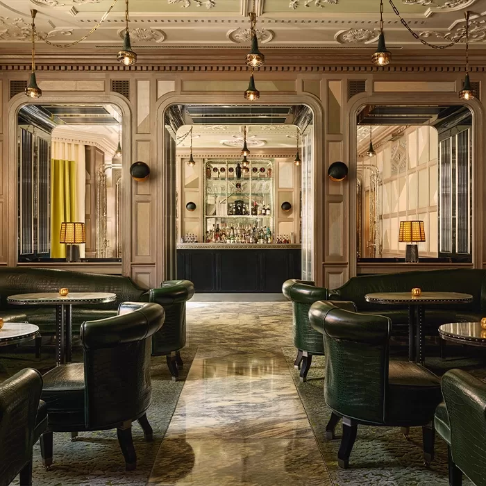 Elegant lounge with dark green leather chairs, round tables, ornate ceiling, hanging lights, and a bar with mirrored shelves in the background. The space has a luxurious, vintage ambiance.