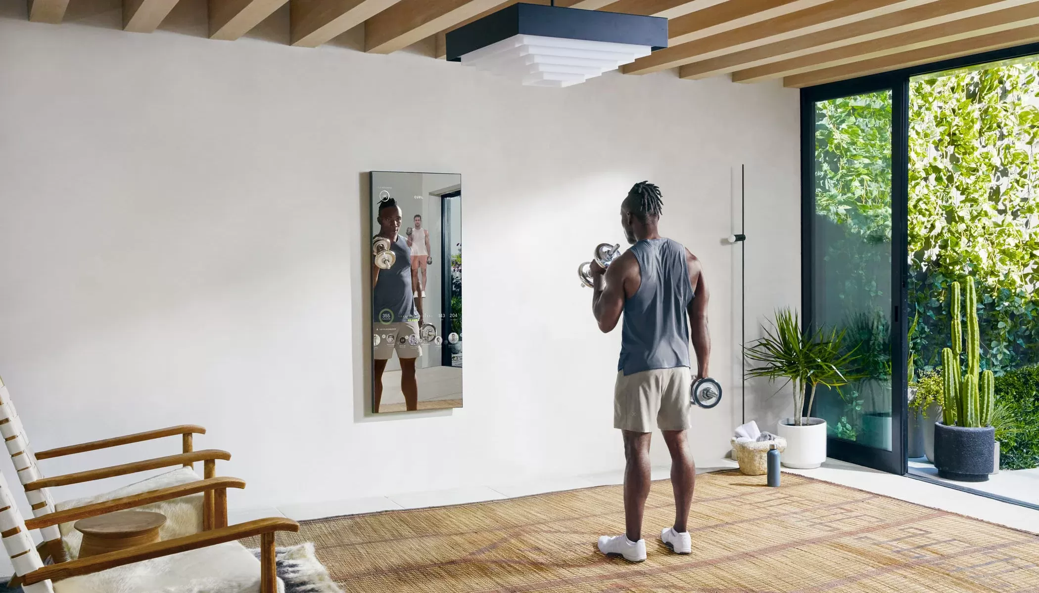 Man in gym clothes with weights working out opposite a home fitness technology mirror in a sunny room