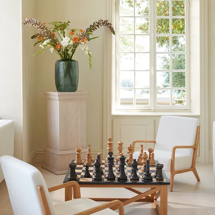 A sunlit room inside Domaine de Bagatelle features a chess table with large black and brown pieces. Two white armchairs surround the table. A tall vase with a floral arrangement stands on a pedestal by a multi-paned window with a view of greenery outside.