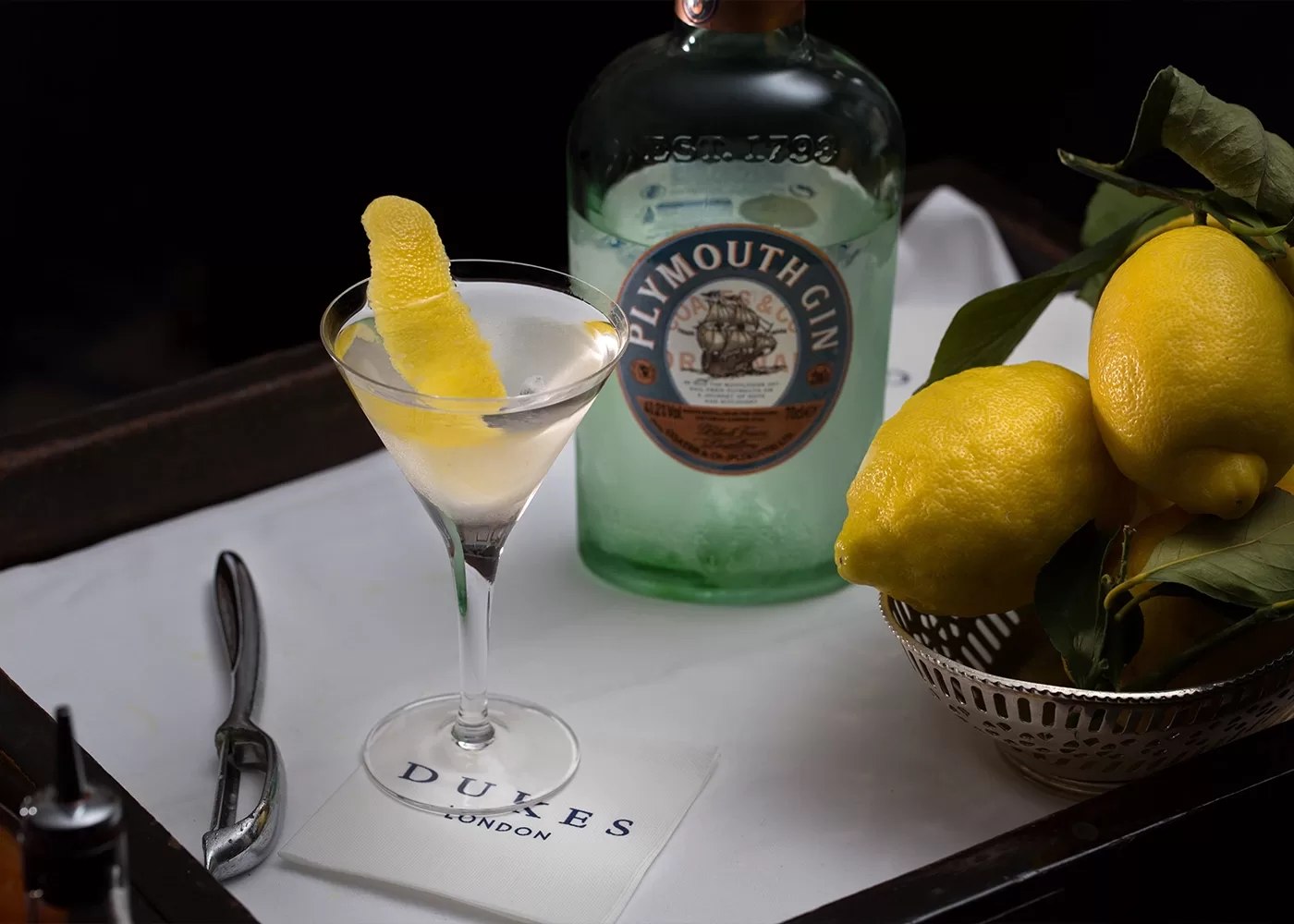 A martini with a lemon twist sits on a tray beside a bottle of Plymouth Gin, fresh lemons in a bowl, a napkin labeled DUKES LONDON, and a cocktail spoon.