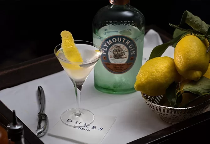 A martini with a lemon twist sits on a napkin labeled DUKES LONDON, next to a bottle of Plymouth Gin, a bar spoon, and a metal bowl filled with fresh lemons.