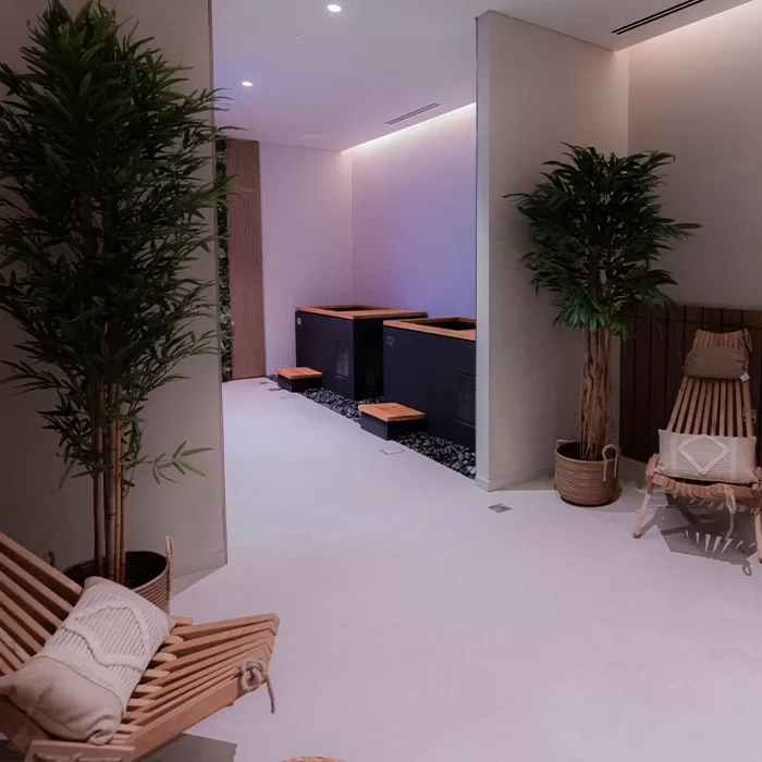 A serene spa room with wooden lounge chairs, potted green plants, and three dark soaking tubs against the back wall, softly lit with ambient lighting for a relaxing atmosphere.