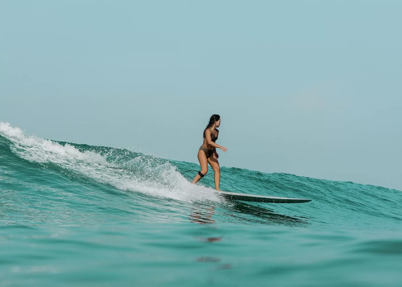 A woman in a swimsuit is surfing on a wave in the ocean. She is standing on a surfboard, riding along a crest with water splashing around. The sky is clear, adding to the serene and adventurous atmosphere.