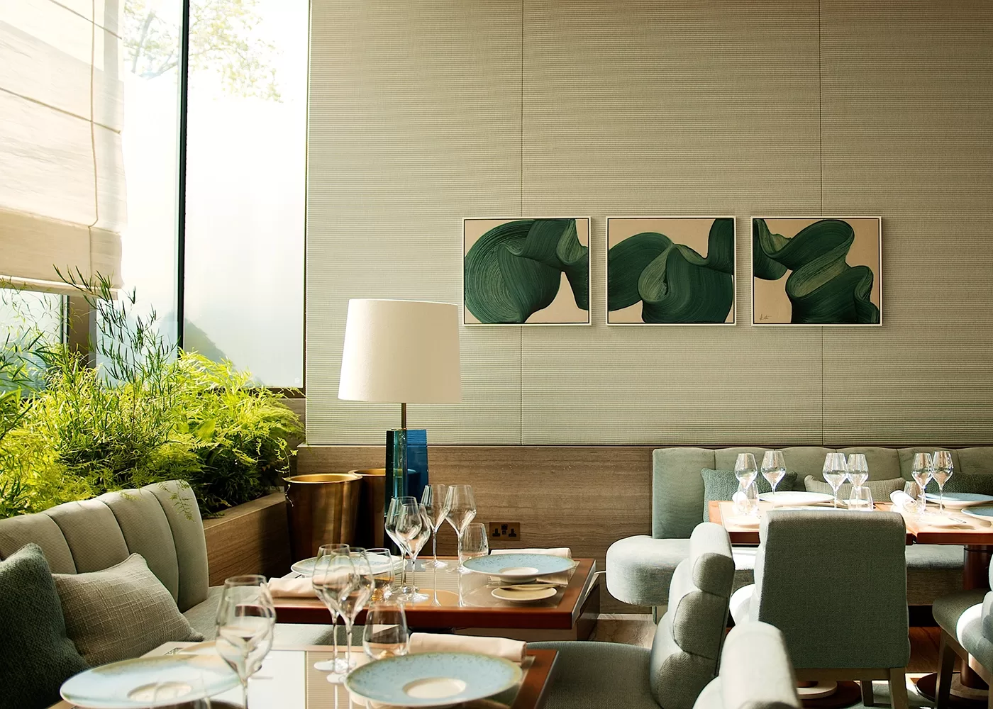 A modern dining room with green cushioned chairs and wooden tables set with glassware and blue-rimmed plates. A trio of abstract green artworks hang on the wall, and a large window lets in natural light, illuminating a potted plant nearby.
