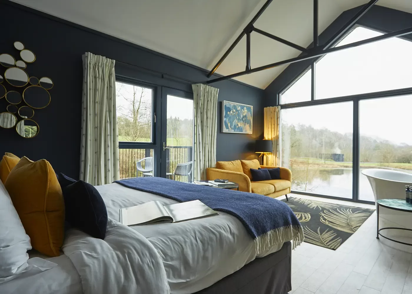 A modern bedroom with large windows overlooks a scenic outdoor landscape. The room features a bed with a blue blanket, a yellow sofa, circular mirrors on the wall, and a comfortable chair. Natural light fills the space, creating a cosy atmosphere.