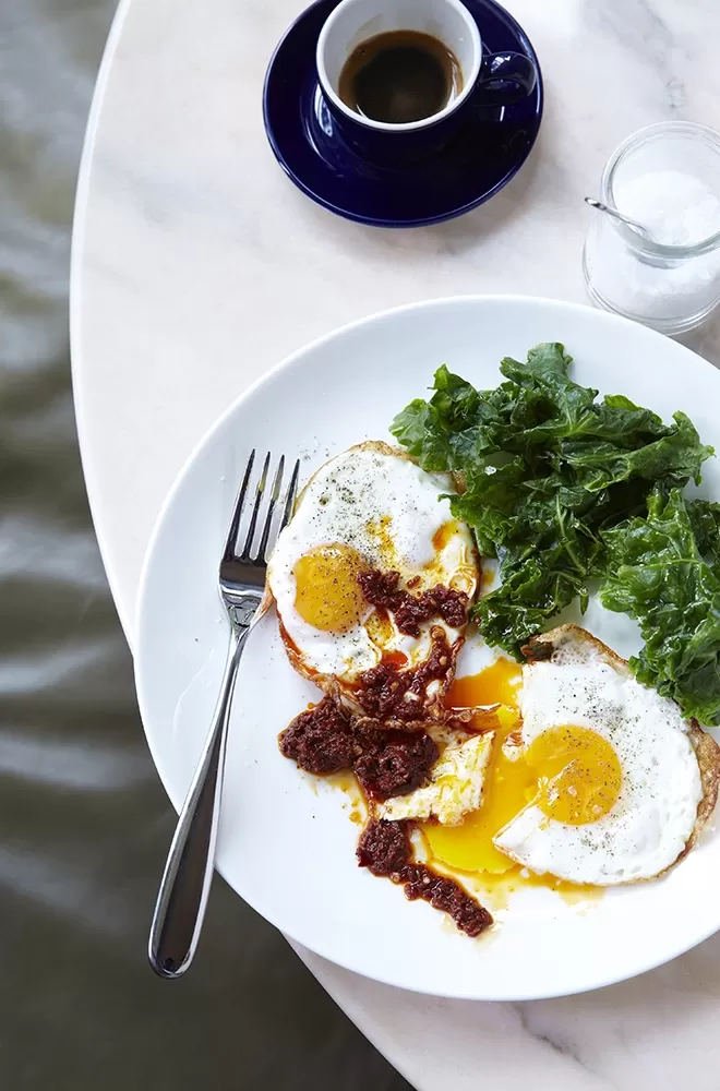 A breakfast plate with two sunny-side-up eggs, greens, and a mound of sauce on a white plate, next to a fork. A small cup of coffee and a glass of cream are on the table, which has a marble-like surface
at Granger & Co, London.