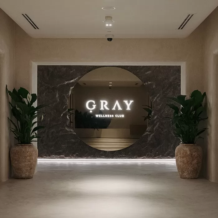 A modern hallway with two large potted plants leads to a circular entrance framed in dark stone, featuring an illuminated sign that reads GRAY WELLNESS CLUB.