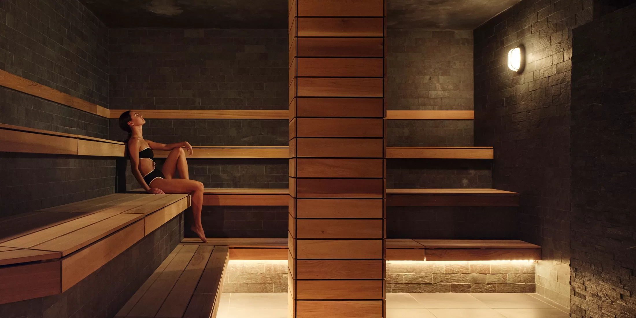 A person relaxes in a dimly lit sauna, sitting on a wooden bench with legs stretched out. The room features dark stone walls, wooden benches, and a central wooden pillar. Soft lighting creates a warm ambiance.