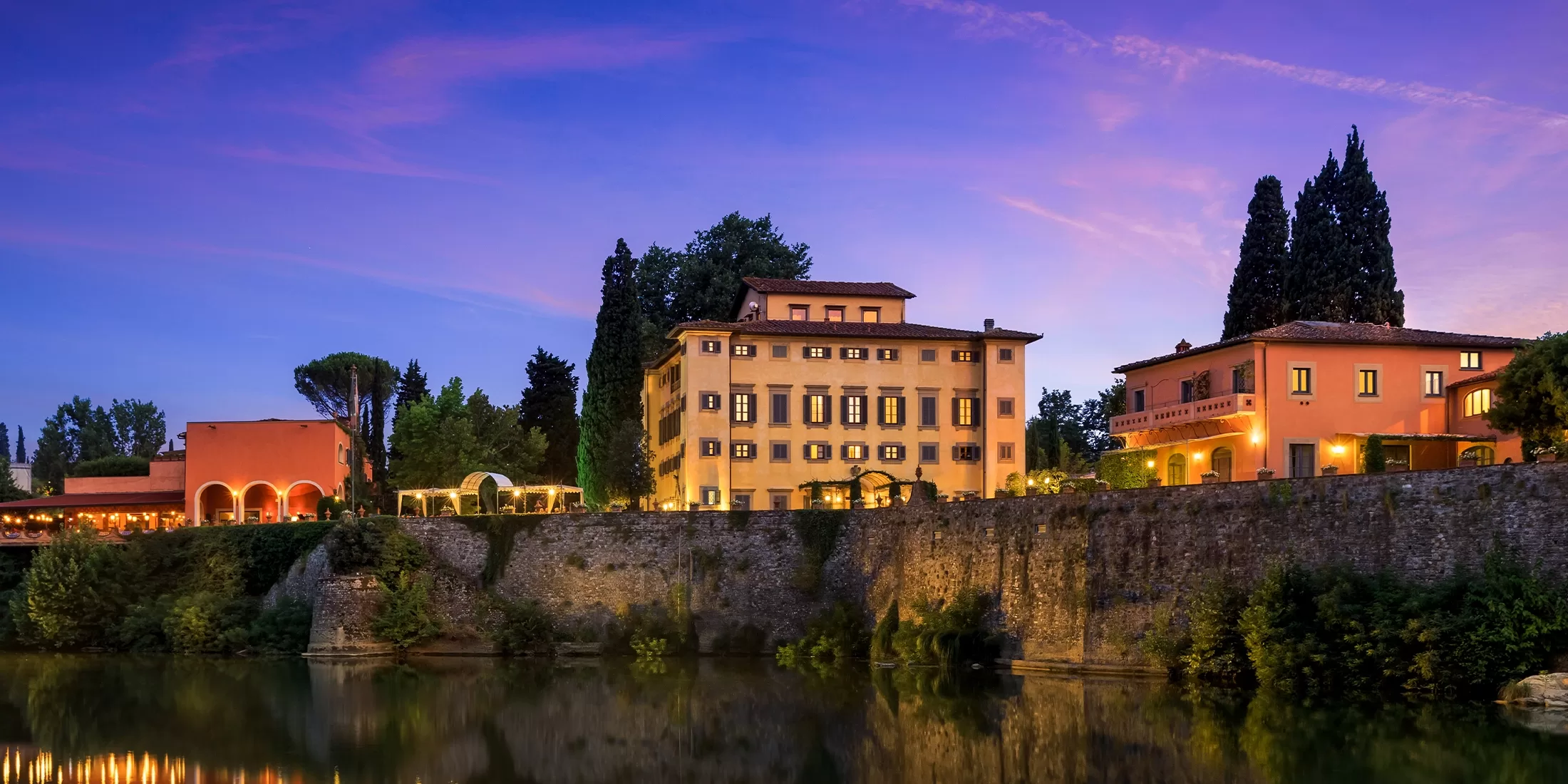 A row of elegant, illuminated buildings with terracotta roofs sits atop a stone embankment, reflected in a calm river at dusk under a purple and pink sky, surrounded by tall trees.