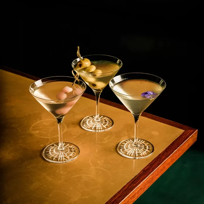 Three martini glasses with clear cocktails sit on a golden table; one is garnished with cocktail onions, one with green olives, and one with a purple flower.