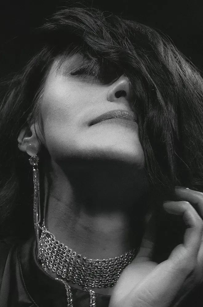 Black and white photo of a woman wearing diamond jewellery; a choker necklace and longline earrings posing with her hair covering her face.