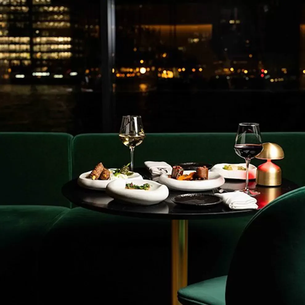 Food and drink served on round dining table with green velour sofa interior at Hithe + Seek, London