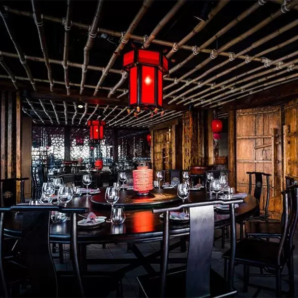 A dimly lit restaurant with a round table set for a meal, featuring neatly arranged glasses and napkins. Red lanterns hang from a bamboo ceiling, casting a warm glow. The decor is rustic with wooden walls and traditional Asian elements at Hutong: The Shanghai Room.