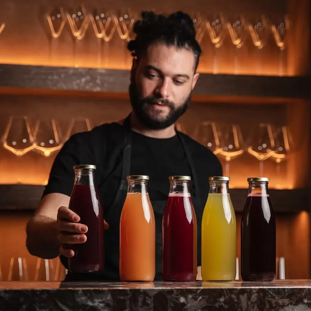 A person stands behind a marble counter at Akoko, Fitzrovia, arranging five bottles filled with colorful liquids. The background features rows of glassware on illuminated shelves. The person has dark hair tied up and is wearing a black shirt with an apron.