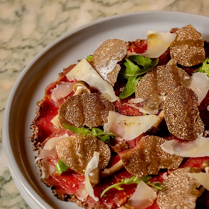 A close-up of a gourmet dish featuring thin slices of beef carpaccio, topped with fresh arugula, shaved Parmesan cheese, and generous slices of black truffle on a white plate.