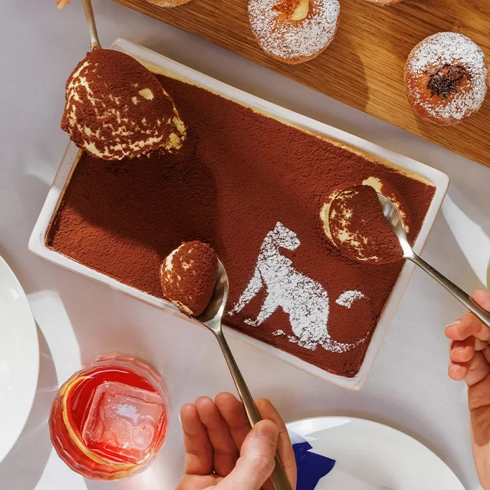 A rectangular tiramisu dessert dusted with cocoa powder, featuring a powdered sugar outline of a cat. Two hands are scooping portions with spoons. Other desserts and a red drink are nearby on the table.