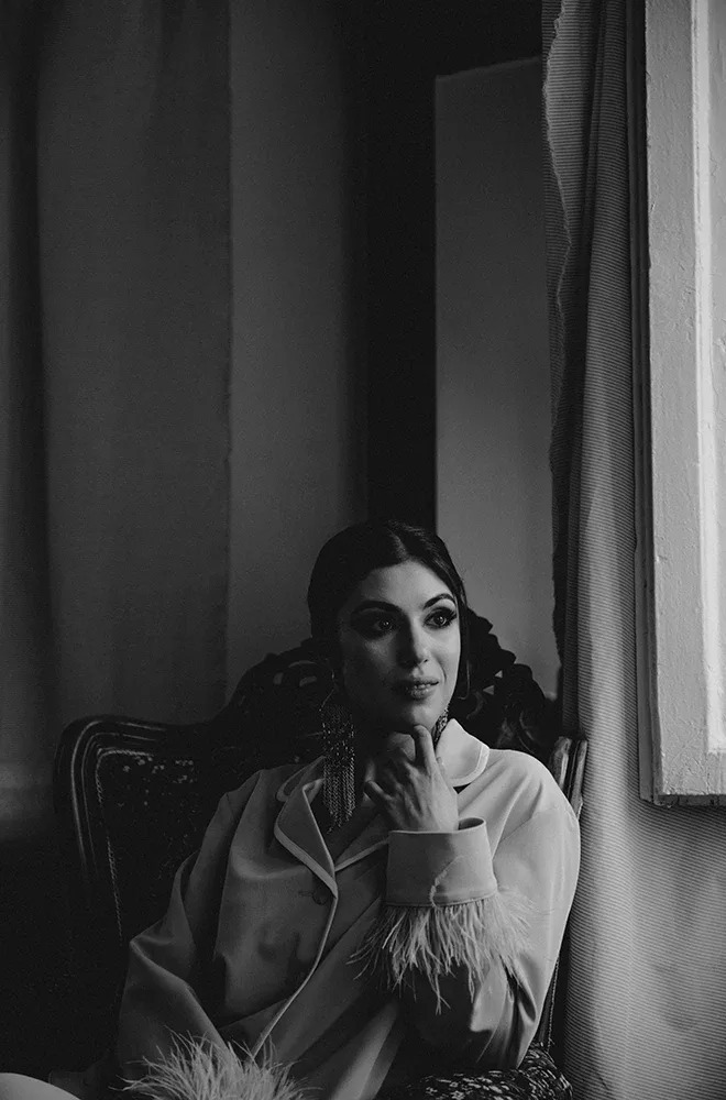 Black and wite photo of a glamorous woman wearing pyjamas with feather sleeves wearing longline statement earrings, sitting on a chair and looking out of a window.