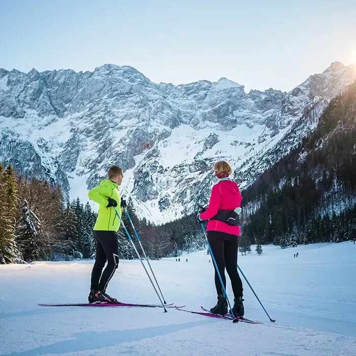 Two people in bright winter jackets cross-country skiing on snowy terrain, surrounded by trees and tall, snow-covered mountains under a clear sky with sunlight peeking over the peaks.