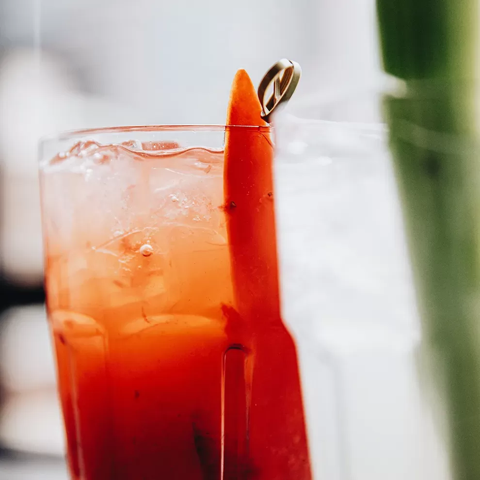 Close-up of a glass filled with a bright orange-red iced beverage, garnished with a slice of vegetable and a green leaf. The glass is adorned with a gold ring on the rim, adding a decorative touch. Served at Mauro Colagreco at Raffles London at The OWO, Whitehall.