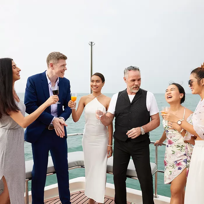 Six people dressed in elegant summer outfits stand on a yacht, smiling and holding drinks, enjoying a sunny day over the water. The background shows calm sea and a clear sky.