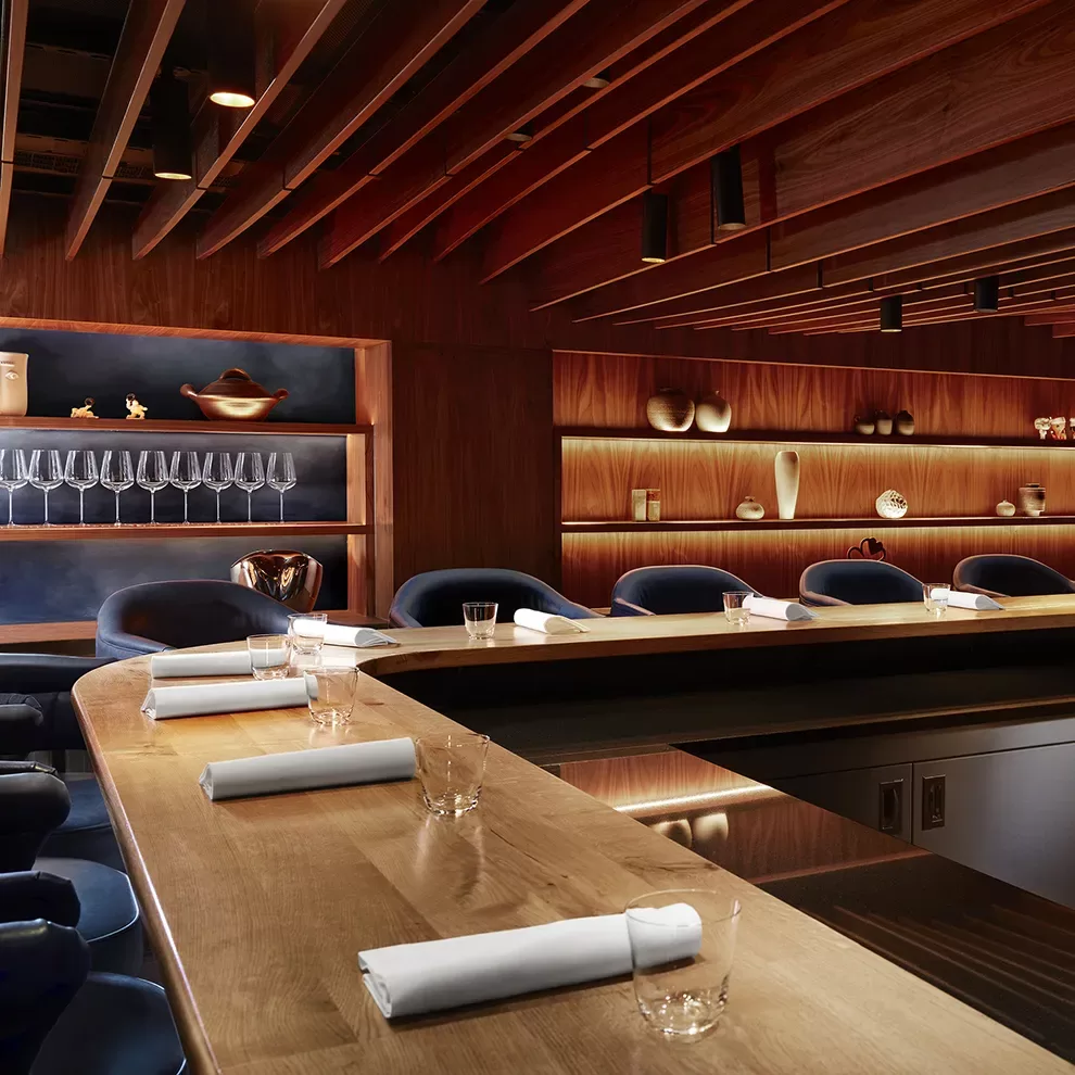 The modern, elegant restaurant interior of Kitchen Table, Fitzrovia, with a long, wooden counter surrounded by navy blue chairs. Soft, warm lighting highlights glassware and decorative items on wooden shelves, creating a cosy, sophisticated ambiance.